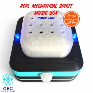 Puede incluir: Caja de música mecánica con el texto "REAL MECHANICAL SPIRIT MUSIC BOX SWAN LAKE". La caja es negra con un borde azul claro y una parte superior blanca con varios agujeros. Tiene luces azules a cada lado.