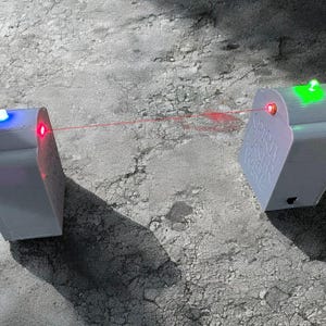 May include: Two white rectangular electronic devices with rounded tops, connected by a thin red laser beam. The left device features a blue light on top, while the right device displays a green light on top and the embossed text SPIRIT LASER WIRE.