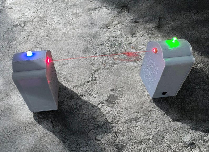 May include: Two white rectangular electronic devices with rounded tops, connected by a thin red laser beam. The left device features a blue light on top, while the right device displays a green light on top and the embossed text SPIRIT LASER WIRE.
