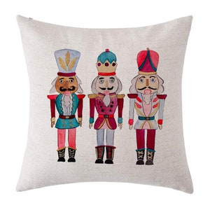 Op de afbeelding: Drie aquarel-notenkrakers op een witte linnen kussenhoes. De notenkrakers dragen rode, blauwe en groene uniformen met gouden accenten. Ze hebben witte baarden en snorren.