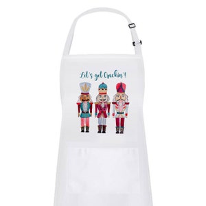 May include: White kitchen apron with a pocket, featuring three nutcracker figurines and the text "Let's get Crackin'!".