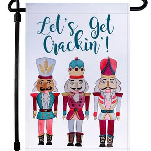 May include: A white garden flag with three watercolor nutcracker illustrations. The text "Let's Get Crackin'" is written in teal blue lettering.