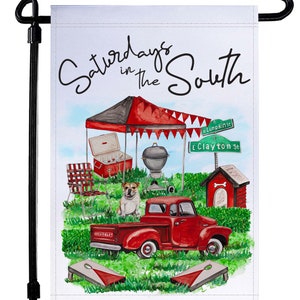May include: A white garden flag with a watercolor illustration of a red pickup truck, a red and white tent, a grill, a cooler, a dog house, and two cornhole boards. The text "Saturdays in the South" is written in black cursive at the top of the flag.