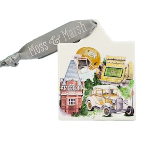 May include: A white ceramic ornament with a yellow and white Georgia Tech logo, a yellow and white football helmet, a yellow and white football stadium, a red brick building with the words "Tech" on the front, and a yellow and white vintage car. The ornament has a gray ribbon with the words "Moss & Marsh" on it.