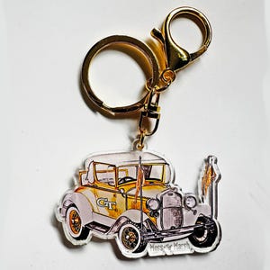 May include: A gold-colored keychain with a vintage car design. The car is primarily white and yellow with black accents and the letters "GT". The keychain has a gold ring and clasp. The text "Moss for Marsh" is at the bottom.