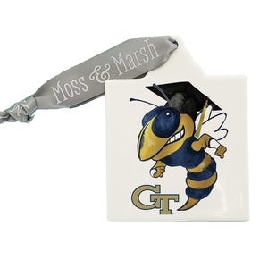 May include: A white ceramic ornament with a cartoon bee wearing a graduation cap and holding a diploma. The bee is yellow and blue with gold accents. The ornament has the letters "GT" in gold on the bottom. The ornament is attached to a gray ribbon with the text "Moss & Marsh" printed on it.