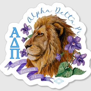 May include: A watercolor illustration of a lion's head with purple violets and green leaves. The lion is facing left and has a serious expression. The text "Alpha Delta Pi" is written in a cursive font around the lion's head. The text "We live for each other est 1851" is written on a purple ribbon below the lion's head. The letters "AΔΠ" are written in blue on the left side of the image.