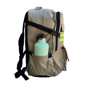 Puede incluir: Una mochila beige con correas negras y una franja reflectante verde. Una botella de agua azul claro est&aacute; en el bolsillo lateral.