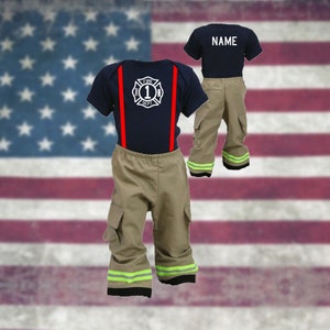 May include: A firefighter costume for a baby or toddler, featuring a navy blue shirt with a red trim and a white firefighter emblem, beige pants with green reflective stripes, and red suspenders. The shirt has the text "FIRE DEPT 1" on it. The pants have a cargo pocket on the left side. The costume is displayed against a backdrop of an American flag.