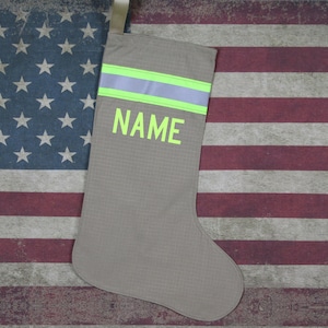 May include: A gray stocking with a green and white reflective stripe and the word "NAME" in green letters. The stocking is on a red, white, and blue American flag background.