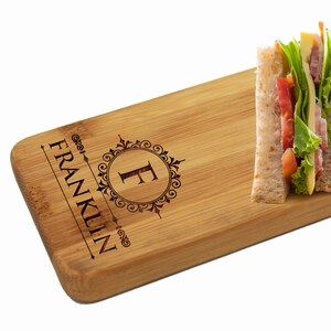 May include: A personalized bamboo serving board with a decorative monogram and the text "FRANKLIN" engraved on the surface. The board features a long handle and is displayed with four sandwiches on top.