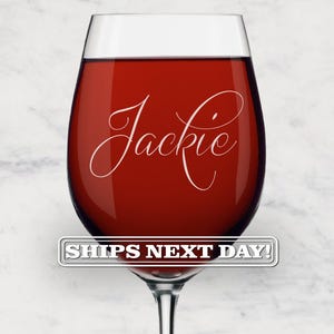 May include: A clear glass wine glass filled with deep red liquid. The glass features the name Jackie written in a white cursive script font across the center. A rectangular label with a silver border and white text reads SHIPS NEXT DAY!