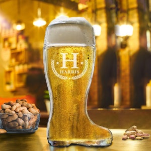 May include: A personalized beer boot glass with a white letter H and the name HARRIS etched on the front. The glass is filled with beer and has a white foam head. The glass is sitting on a wooden table with peanuts in a bowl next to it.