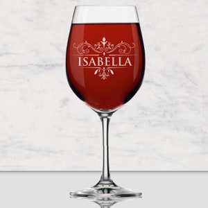 May include: A clear glass wine glass filled with deep red wine. The glass features a white etched design with decorative scrollwork and the name ISABELLA centered in capital letters. The stem and base are transparent glass with a smooth finish.