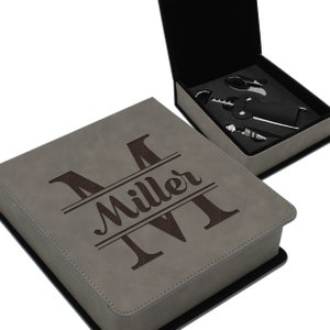 May include: A gray faux leather box with a black trim and a brown monogrammed initial "M" with the name "Miller" underneath. The box is open to reveal a set of wine tools.