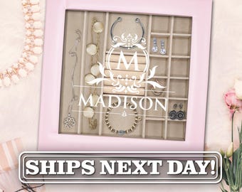 May include: A square jewellery organiser box with a light pink frame and a glass lid featuring a white printed monogram with the letter M and the name MADISON. The interior is divided into beige compartments containing various necklaces, earrings, and bracelets.