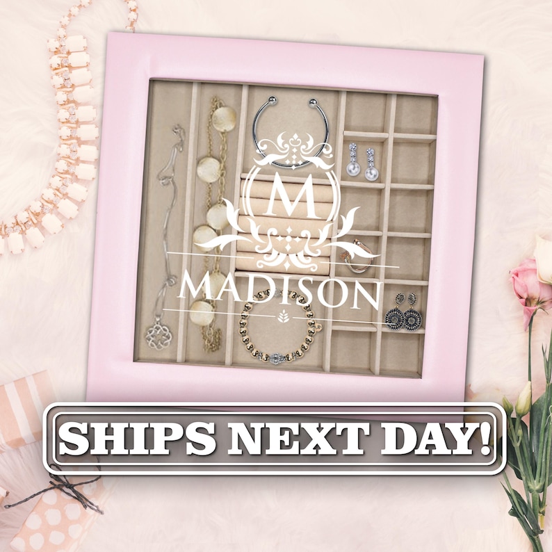 May include: A square jewelry organizer box with a light pink frame and a glass lid featuring a white printed monogram with the letter M and the name MADISON. The interior is divided into beige compartments containing various necklaces, earrings, and bracelets.