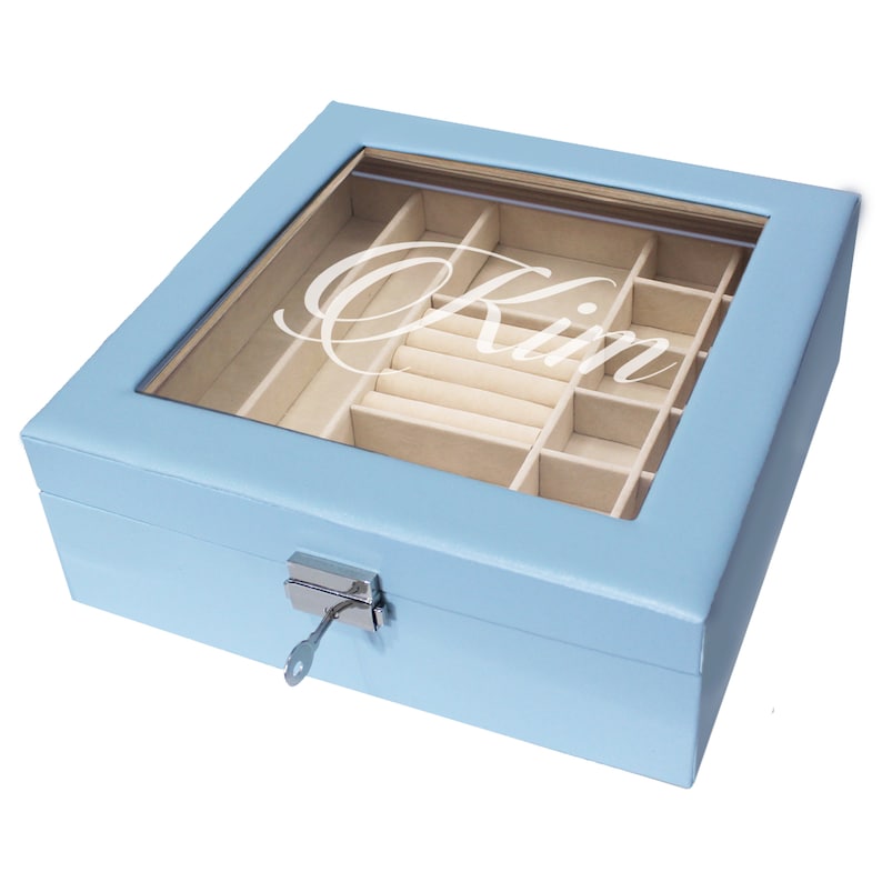 May include: A light blue jewelry box with a clear glass lid featuring the white script text Kim. The box includes a beige interior with multiple compartments and a ring holder section. A silver metal latch with an inserted key is on the front.
