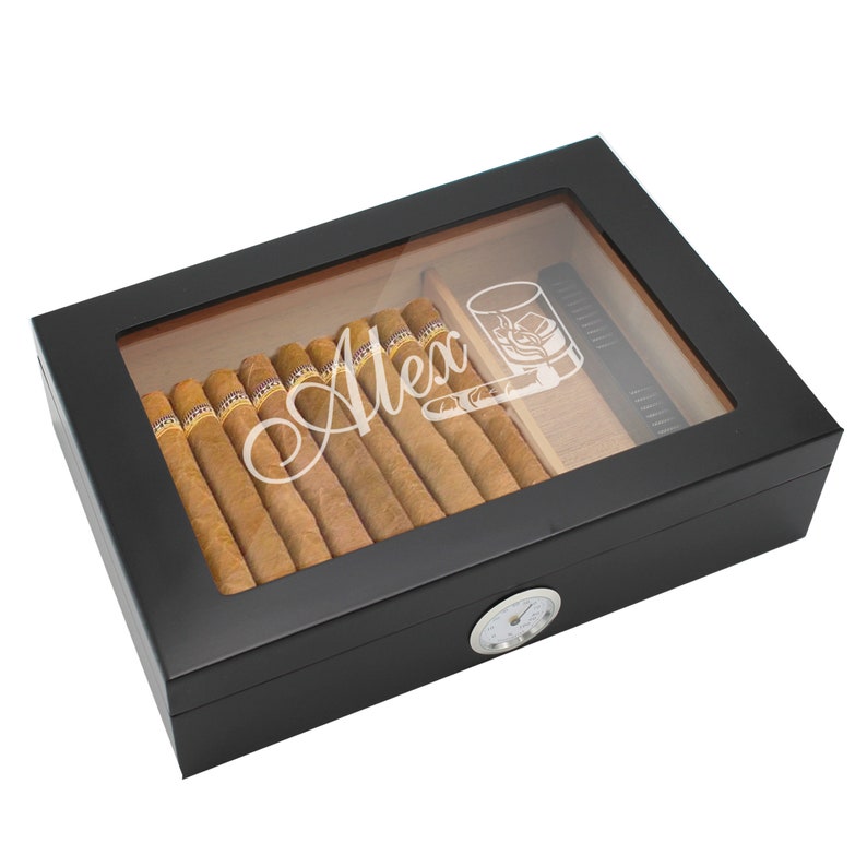 May include: A black rectangular humidor with a glass top featuring the white engraved name Alex and a graphic of a glass with ice. Inside are several brown cigars and a black rectangular accessory. A round silver analog hygrometer is on the front.