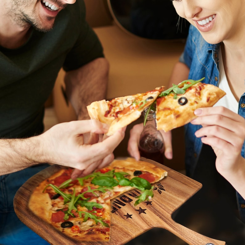 Peut inclure: Une pelle &agrave; pizza en bois avec une pizza garnie de pepperoni, d'olives et de roquette. Deux personnes partagent la pizza. La pelle a une poign&eacute;e et le mot "INDULGE" est visible sur la pelle.