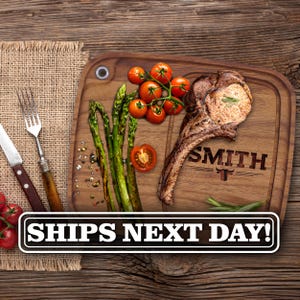 May include: A rectangular wooden cutting board featuring a metal hanging hole, engraved with the name SMITH and a bull icon. The board is topped with a cooked bone-in steak, fresh green asparagus spears, and cherry tomatoes on the vine.