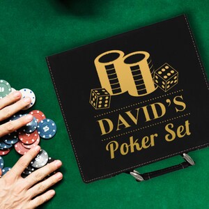 Puede incluir: Malet&iacute;n negro para juego de p&oacute;quer con gr&aacute;ficos dorados de dados y fichas, y el texto "DAVID'S Poker Set". El malet&iacute;n est&aacute; sobre una superficie de fieltro verde con fichas de p&oacute;quer esparcidas.