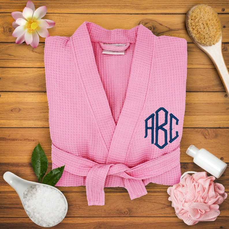 May include: Pink waffle weave bathrobe with a navy blue monogram "ABC" embroidered on the front. The robe has a sash tie and is laid out on a wooden surface.