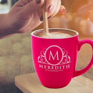 May include: A vibrant pink coffee mug with a white monogram "M" and the name "Meredith" printed on the front. The mug is filled with coffee, and a wooden spoon stirs the contents. The mug sits on a wooden surface.