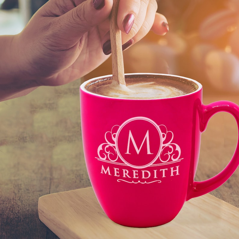 May include: A vibrant pink coffee mug with a white monogram "M" and the name "Meredith" printed on the front. The mug is filled with coffee, and a wooden spoon stirs the contents. The mug sits on a wooden surface.
