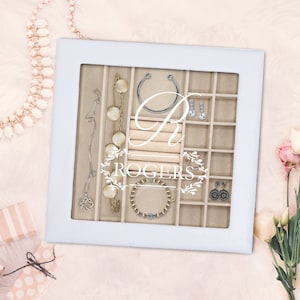 May include: A white square jewelry display case with a glass lid featuring the white printed text R ROGERS. The interior is lined with beige fabric and divided into multiple compartments containing various necklaces, earrings, and bracelets.
