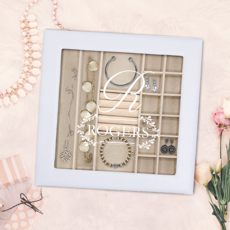 May include: A white square jewelry display case with a glass lid featuring the white printed text R ROGERS. The interior is lined with beige fabric and divided into multiple compartments containing various necklaces, earrings, and bracelets.