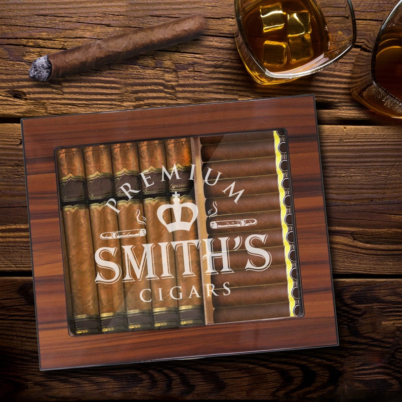 May include: A rectangular wooden cigar humidor with a glass top featuring the white printed text PREMIUM SMITH'S CIGARS and a crown icon. The humidor is filled with rows of brown cigars, with a single cigar and a glass of amber liquid nearby.