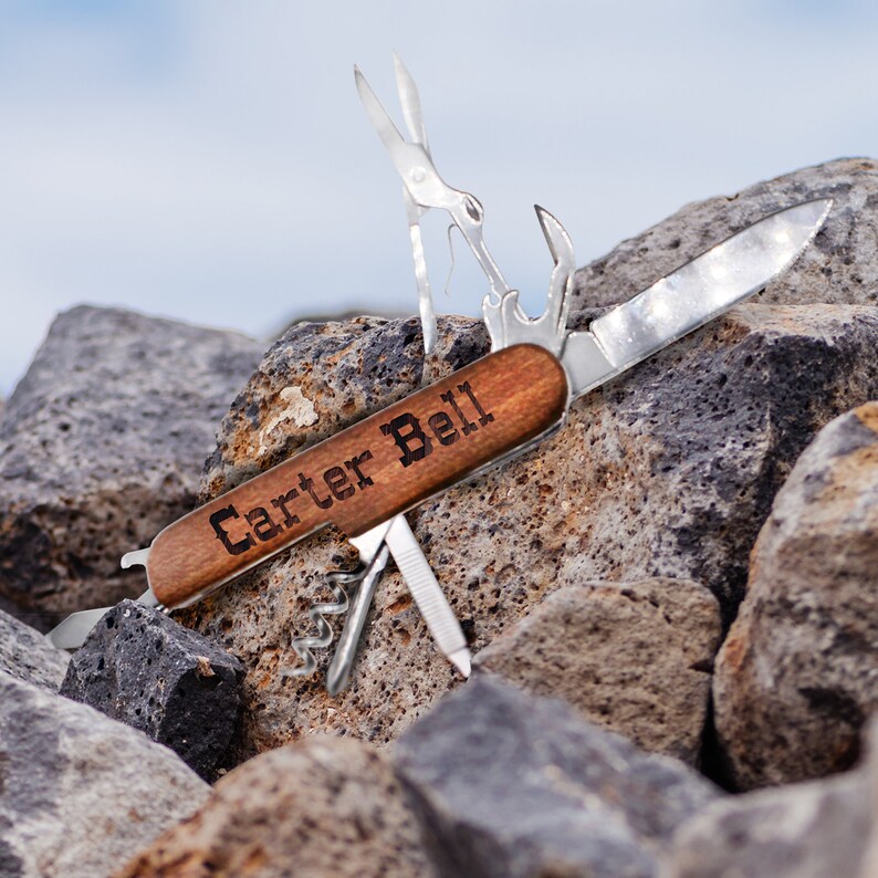 Puede incluir: Una navaja multiusos de madera marr&oacute;n con una hoja de plata y un abridor de botellas. El mango est&aacute; grabado con el nombre "Carter Bell".
