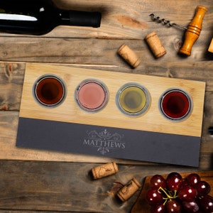 May include: A bamboo and slate wine flight set with four glass tasting cups, each filled with a different colored wine. The slate base is engraved with the name "MATTHEWS". Corks, a wine bottle, and a corkscrew are also visible.