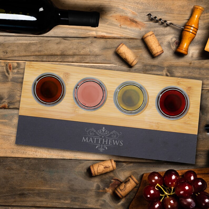 May include: A bamboo and slate wine flight set with four glass tasting cups, each filled with a different colored wine. The slate base is engraved with the name "MATTHEWS". Corks, a wine bottle, and a corkscrew are also visible.