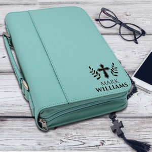May include: A light teal Bible cover with a zipper closure and a handle. The cover features a black cross design with the name "MARK WILLIAMS" printed below. The cover is made of a smooth material.