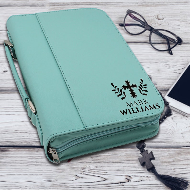 May include: A light teal Bible cover with a zipper closure and a handle. The cover features a black cross design with the name "MARK WILLIAMS" printed below. The cover is made of a smooth material.