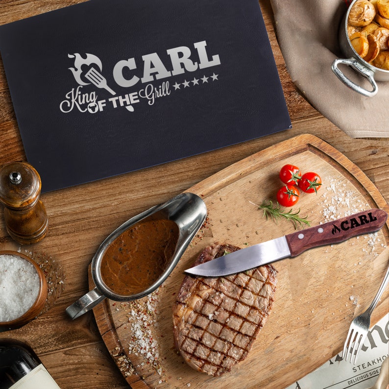 May include: A wooden cutting board with a steak, a gravy boat filled with brown gravy, a wooden handled knife with the text "CARL" on the handle, and a small plate of salt. The cutting board is on a wooden table with a black placemat with the text "CARL King of the Grill" on it.