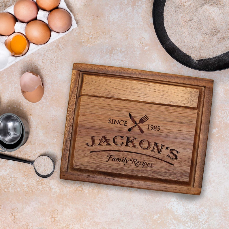 Puede incluir: Una tabla de cortar rectangular de madera con un canal para jugos, grabada con el texto "SINCE 1985", "JACKON'S" y "Family Recipes" junto a un icono de un tenedor y un cuchillo cruzados. La tabla presenta un acabado de veta de madera natural.