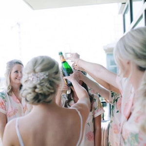 May include: A group of people in floral print robes raise champagne glasses and a bottle of champagne in celebration. The person in the center is wearing a white dress.