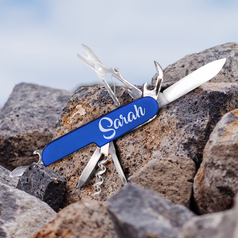 Puede incluir: Un cuchillo multiusos de bolsillo azul con una inscripci&oacute;n blanca que dice "Sarah". El cuchillo est&aacute; abierto y la hoja es visible. El cuchillo est&aacute; sentado en una pila de rocas.