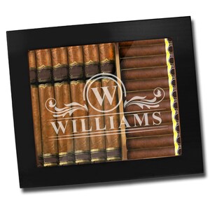 May include: A black rectangular cigar humidor with a glass top featuring a white etched design of the letter W inside a circle, decorative flourishes, and the name WILLIAMS. The interior holds two rows of brown cigars with gold bands.