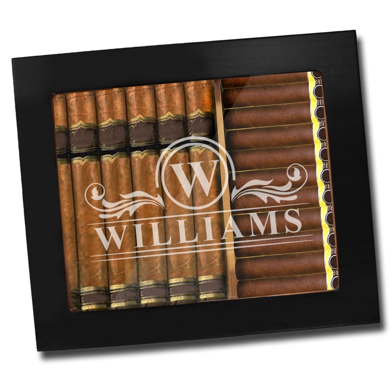 May include: A black rectangular cigar humidor with a glass top featuring a white etched design of the letter W inside a circle, decorative flourishes, and the name WILLIAMS. The interior holds two rows of brown cigars with gold bands.