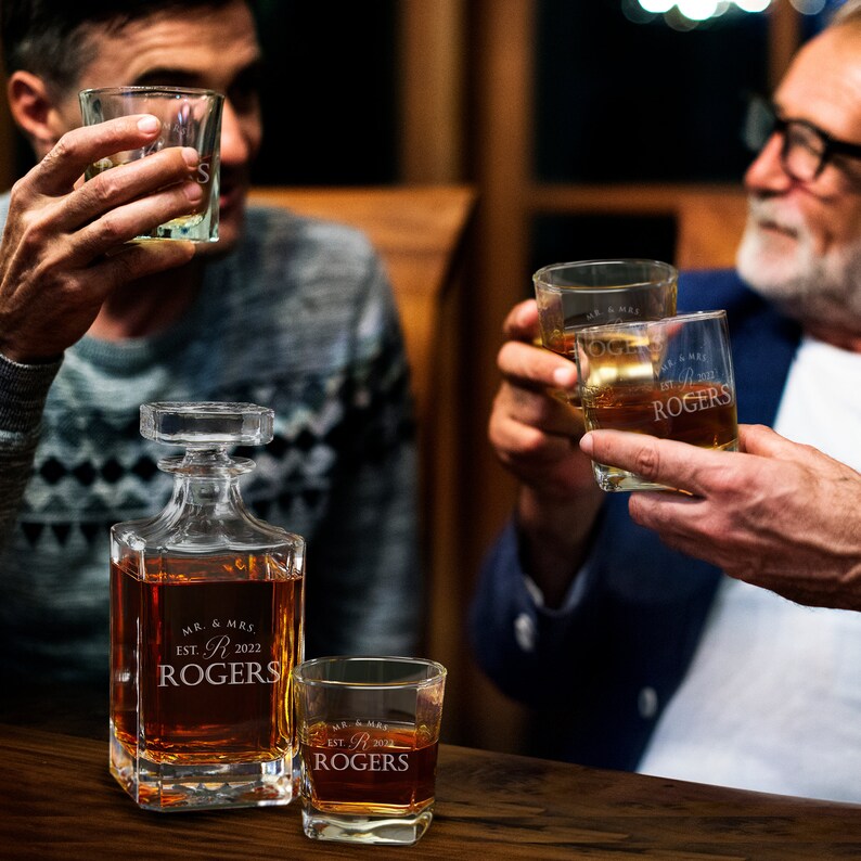 May include: A set of three personalized glass decanters and glasses. The decanter and glasses are engraved with the text "Mr. & Mrs. Rogers Est. 2022".
