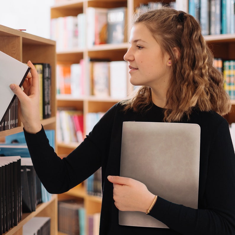 May include: A light grey, rectangular portfolio folder with a smooth, matte finish held in one hand, while the other hand reaches towards a wooden bookshelf filled with various books. The folder is solid-coloured and features clean, stitched edges.