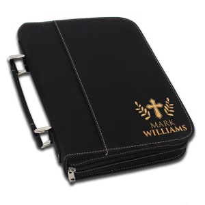 May include: Black leather Bible cover with a handle and zipper closure. Features a gold cross and leaf design with the name "MARK WILLIAMS" printed below. White stitching accents the edges.
