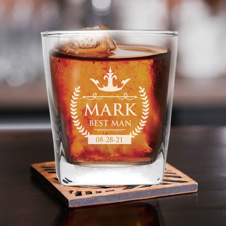 Peut inclure: Un verre &agrave; whisky en verre transparent avec une base carr&eacute;e rempli d'un liquide brun fonc&eacute; et de gla&ccedil;ons. Le verre est d&eacute;cor&eacute; d'un motif blanc avec une couronne, une couronne de laurier et le texte "Mark Best Man 08-28-21".