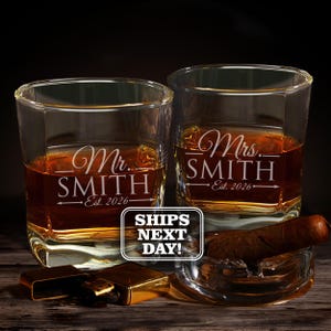 May include: Two clear glass whiskey tumblers filled with amber liquid, each etched with Mr. Smith or Mrs. Smith and Est. 2026. A brown cigar rests in a glass ashtray next to a gold-toned metal lighter. The text SHIPS NEXT DAY is visible.