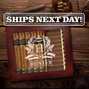 May include: A dark wood cigar humidor with a glass top featuring a white engraved crest with the letter H and the name HUNTER. The humidor is filled with rows of brown cigars, and the glass lid displays the white text SHIPS NEXT DAY!