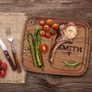 May include: A wooden serving board with a carved groove and the name "SMITH" with a bull graphic. The board holds a grilled steak, asparagus, cherry tomatoes, and rosemary. A knife and fork rest on a burlap cloth.